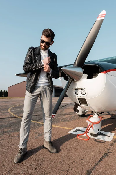 Toute la longueur du pilote élégant en veste en cuir et lunettes de soleil souriant tout en se tenant près de l'avion — Photo de stock