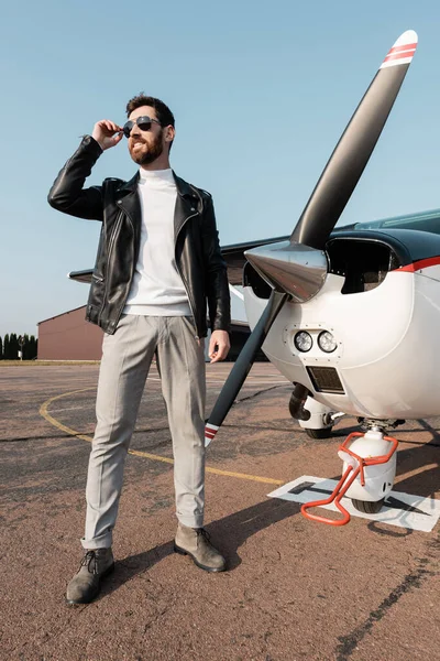 Longitud completa de piloto con estilo en chaqueta de cuero ajustando gafas de sol y sonriendo cerca de aviones - foto de stock