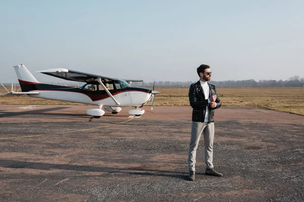 Piena lunghezza di pilota elegante in giacca di pelle e occhiali da sole in piedi vicino all'elicottero — Foto stock