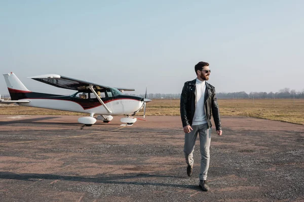 Toute la longueur du pilote élégant en veste en cuir et lunettes de soleil marchant près de l'hélicoptère — Photo de stock