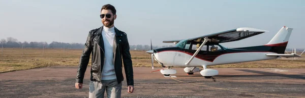 Pilote heureux en veste en cuir élégant et lunettes de soleil marchant près de l'hélicoptère, bannière — Photo de stock