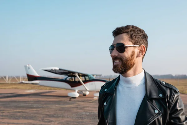 Piloto alegre en elegante chaqueta de cuero y gafas de sol sonriendo cerca de helicóptero borroso - foto de stock