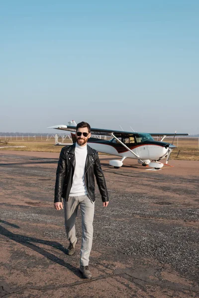 Longitud completa de piloto positivo en elegante chaqueta de cuero y gafas de sol caminando cerca de helicóptero - foto de stock