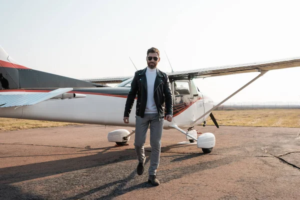 Toute la longueur du pilote en veste en cuir élégant et lunettes de soleil marchant près de l'hélicoptère — Photo de stock