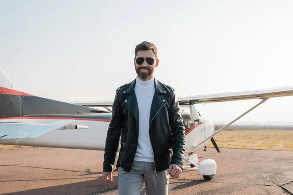 Piloto alegre en elegante chaqueta de cuero y gafas de sol caminando cerca de helicóptero - foto de stock