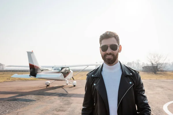 Cheerful pilot in black leather jacket and sunglasses near helicopter — Stock Photo