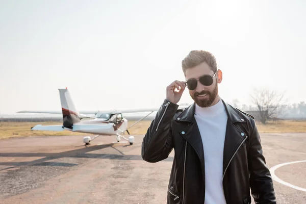 Piloto feliz en chaqueta de cuero negro ajustando gafas de sol cerca de helicóptero - foto de stock