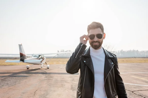 Positive pilot in black leather jacket adjusting sunglasses near helicopter — Stock Photo