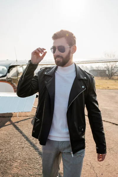 Piloto positivo en chaqueta de cuero y gafas de sol con estilo de pie cerca de helicóptero - foto de stock