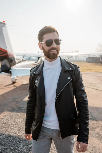 Cheerful pilot in leather jacket and stylish sunglasses standing near helicopter — Stock Photo