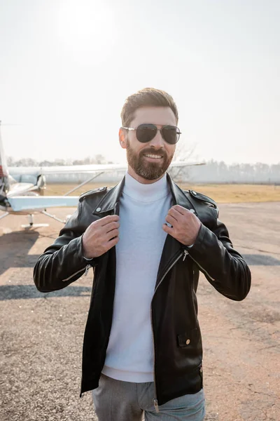 Piloto positivo en gafas de sol con estilo ajustando chaqueta de cuero cerca de helicóptero en el fondo - foto de stock