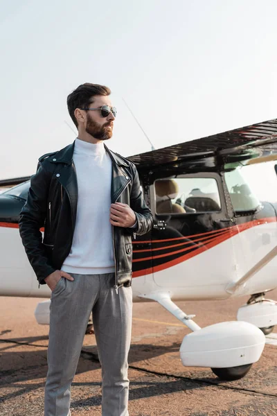 Piloto em óculos de sol elegantes e jaqueta de couro posando com a mão no bolso perto de aeronaves — Fotografia de Stock