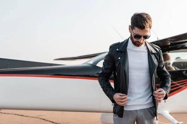 Piloto barbudo en gafas de sol con estilo ajustando chaqueta de cuero cerca de aviones - foto de stock