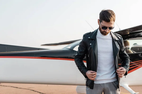 Pilote barbu dans des lunettes de soleil élégantes ajustant veste en cuir près de l'avion — Photo de stock