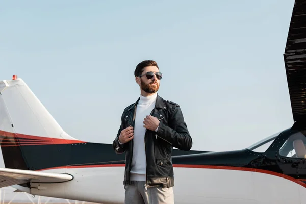 Piloto seguro de sí mismo en gafas de sol elegantes ajustando chaqueta de cuero cerca de aviones - foto de stock