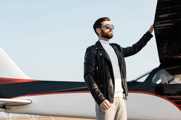 Уверенный пилот в стильных солнцезащитных очках и кожаной куртке стоит рядом с крылом самолета — стоковое фото