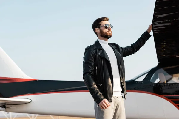 Piloto seguro en gafas de sol con estilo y chaqueta de cuero de pie cerca de ala de avión - foto de stock