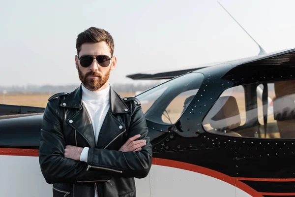 Bearded pilot in stylish sunglasses and leather jacket standing with crossed arms near aircraft — Stock Photo