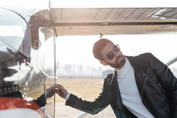 Bearded pilot in sunglasses and leather jacket opening door of helicopter — Stock Photo