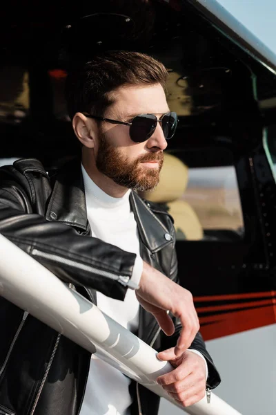 Confident bearded man in sunglasses and black leather jacket near helicopter — Stock Photo