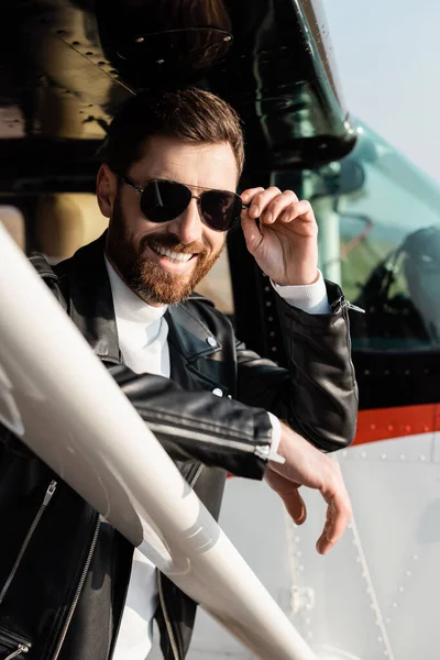 Hombre barbudo feliz ajustando gafas de sol cerca de helicóptero - foto de stock