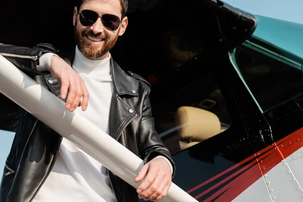 Cheerful bearded man in sunglasses and black leather jacket standing near helicopter — Stock Photo