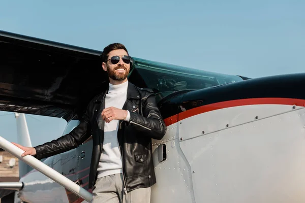 Hombre barbudo feliz en gafas de sol y chaqueta de cuero negro de pie cerca de helicóptero - foto de stock