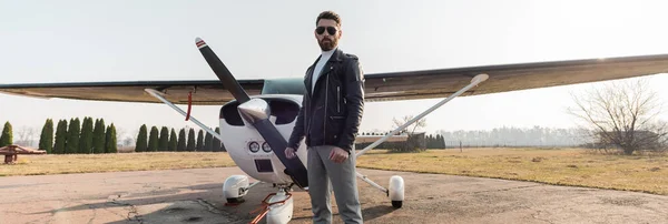 Piloto barbudo en gafas de sol con estilo y chaqueta de cuero de pie cerca de aviones modernos, pancarta - foto de stock