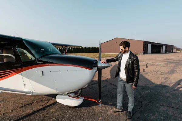 Full length of bearded pilot in stylish sunglasses and leather jacket touching modern aircraft — Stock Photo