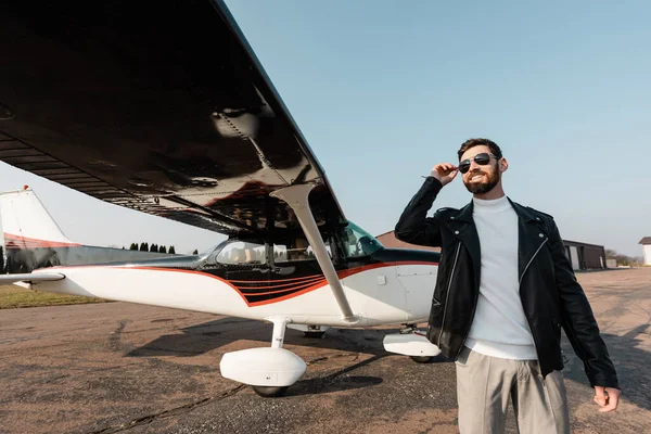 Smiling man in stylish sunglasses and leather jacket standing near modern aircraft — Stock Photo
