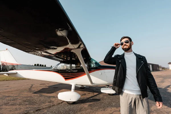 Homme souriant dans des lunettes de soleil élégantes et veste en cuir debout près des avions modernes — Photo de stock