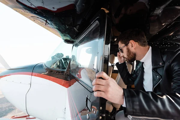 Bärtiger Pilot mit stylischer Sonnenbrille und Lederjacke öffnet Tür im Hubschrauber — Stockfoto