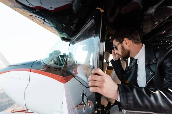 Bearded pilot in stylish sunglasses and leather jacket opening door in helicopter — Stock Photo