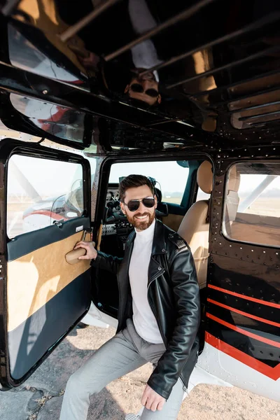 Lächelnder Pilot mit stylischer Sonnenbrille und Lederjacke öffnet Tür im Hubschrauber — Stockfoto