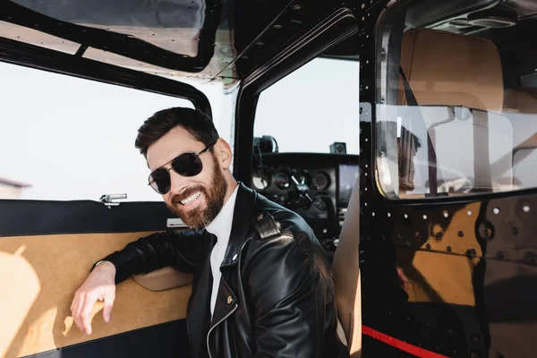 Piloto feliz en gafas de sol con estilo y chaqueta de cuero cerca de la puerta abierta del helicóptero - foto de stock