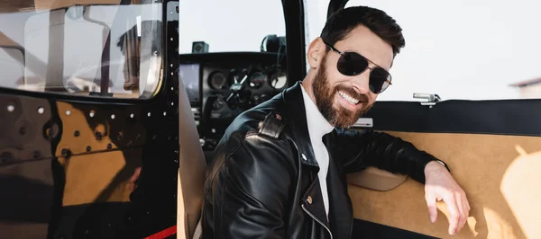 Happy pilot in stylish sunglasses and leather jacket near opened door of helicopter, banner — Stock Photo