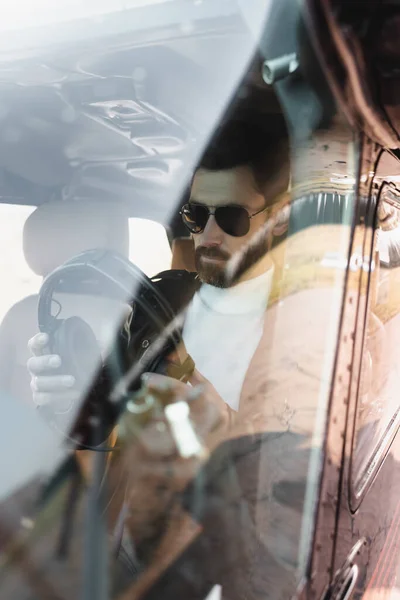 View of bearded pilot in stylish sunglasses holding headphones in cockpit through helicopter windscreen — Stock Photo
