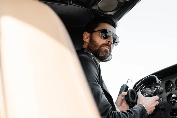 Low angle view of pilot in sunglasses and leather jacket holding headphones in cockpit of helicopter — Stock Photo