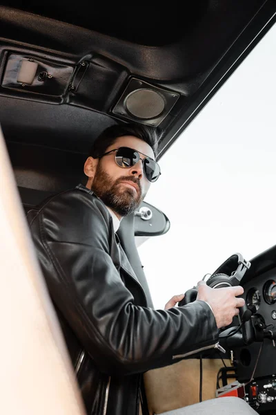 Vista a basso angolo del pilota barbuto in occhiali da sole e giacca di pelle con cuffie in cabina di pilotaggio dell'elicottero — Foto stock