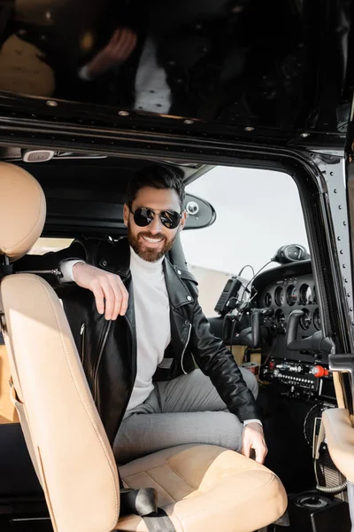 Pilote joyeux dans des lunettes de soleil élégantes et veste en cuir assis dans le cockpit de l'hélicoptère — Photo de stock