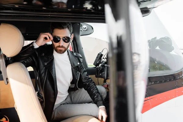 Bearded pilot in leather jacket adjusting stylish sunglasses while sitting in cockpit of helicopter — Stock Photo