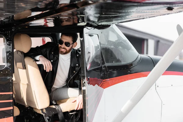 Piloto barbudo en gafas de sol con estilo y chaqueta de cuero sonriendo mientras está sentado en la cabina de helicóptero - foto de stock