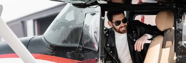 Bearded pilot in stylish sunglasses and leather jacket smiling while sitting in cockpit of helicopter, banner — Stock Photo