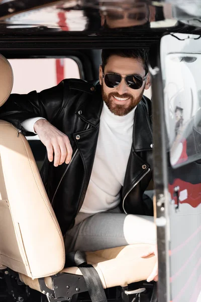 Pilote joyeux dans des lunettes de soleil élégantes et veste en cuir souriant tout en étant assis dans le cockpit de l'hélicoptère — Photo de stock