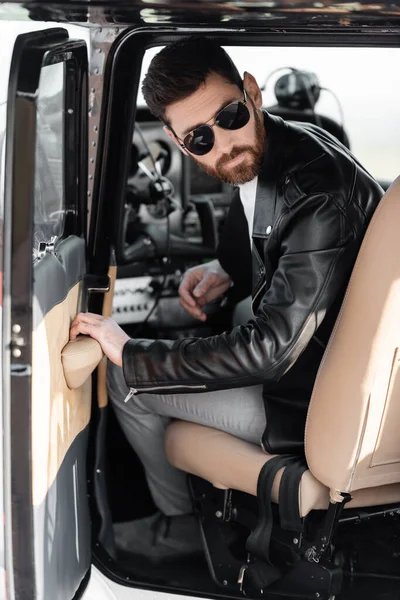 Bearded pilot in stylish sunglasses and leather jacket opening door of helicopter — Stock Photo