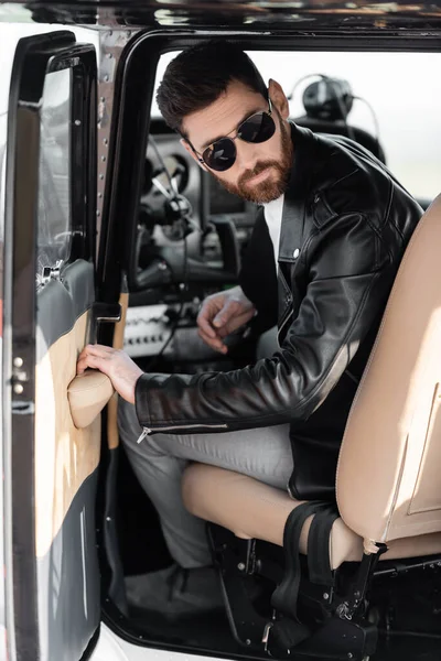 Bärtiger Pilot mit stylischer Sonnenbrille und Lederjacke öffnet Tür des Hubschraubers — Stockfoto