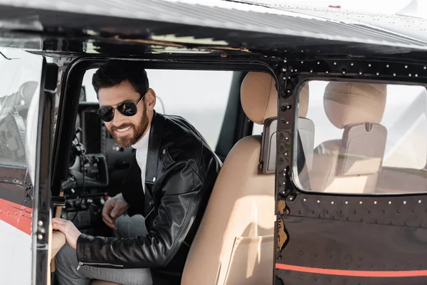 Cheerful pilot in sunglasses and leather jacket opening door while sitting helicopter — Stock Photo