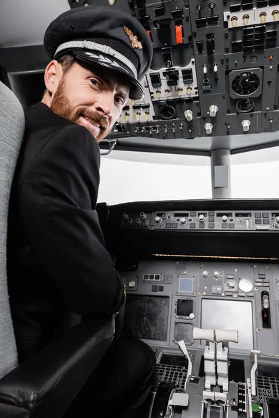Bärtiger Pilot mit Schirmmütze lächelt und blickt in Flugzeugsimulator in die Kamera — Stockfoto