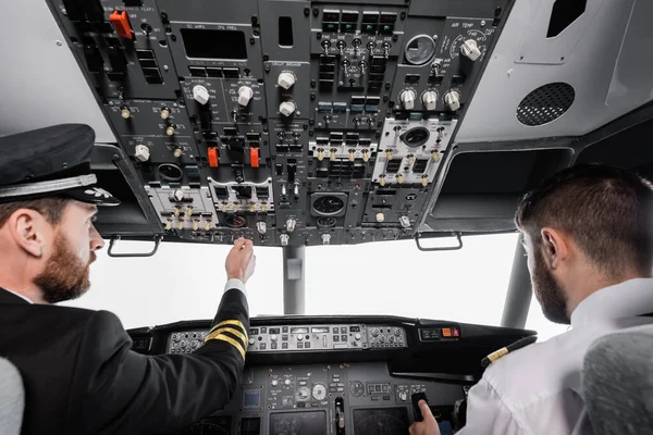 Bearded pilot in cap reaching overhead panel near co-pilot in airplane simulator — Stock Photo