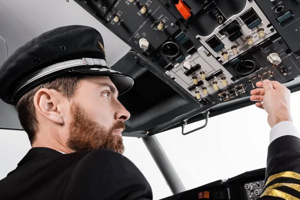 Pilote barbu au capuchon atteignant le panneau supérieur avec un ensemble d'interrupteurs dans le simulateur d'avion — Photo de stock